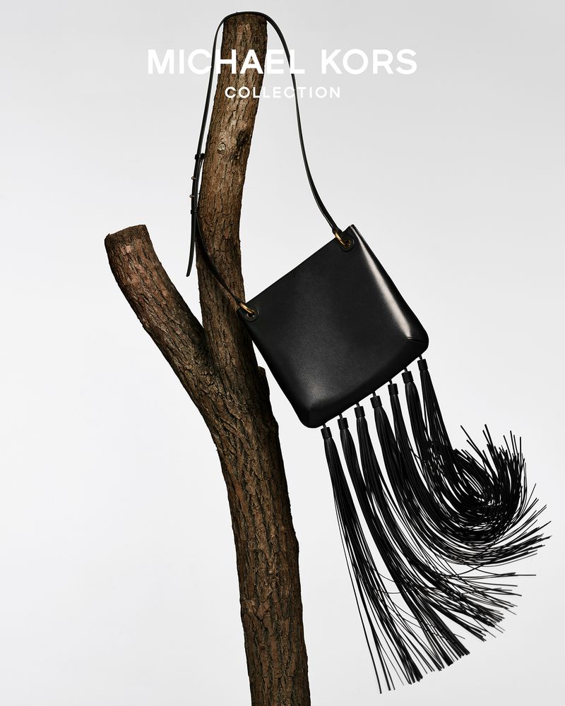 Michael Kors black leather handbag with fringe hanging from a bare tree branch, photographed against a pale gradient backdrop.