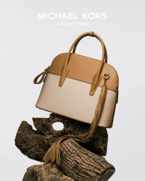 A two-toned leather handbag with a tassel resting on a stack of gnarled wood logs against a white studio background.