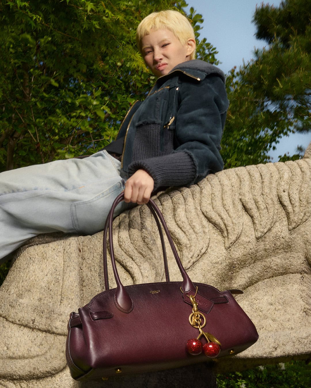 Woman in a bomber jacket poses with a burgundy leather handbag with cherry charm, sitting on a carved tiger in a leafy outdoor setting.
