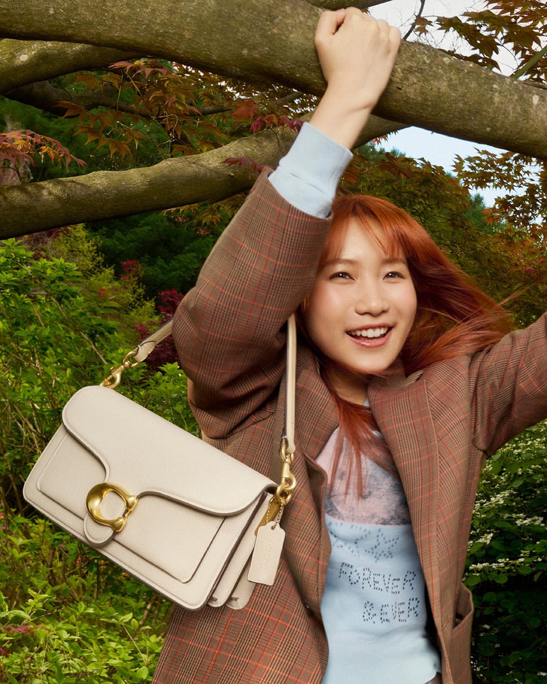 Smiling woman with red hair in a plaid jacket stands near a tree in a park, carrying a beige leather purse on her shoulder.