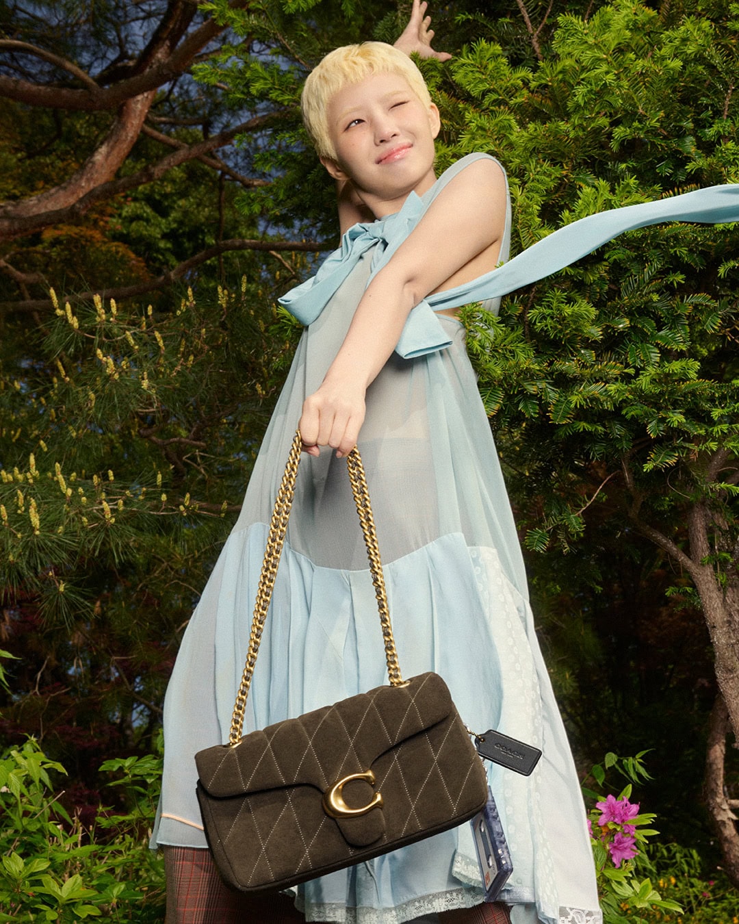 A young adult poses with a brown quilted Coach purse with gold chain strap in a natural outdoor setting with green trees.