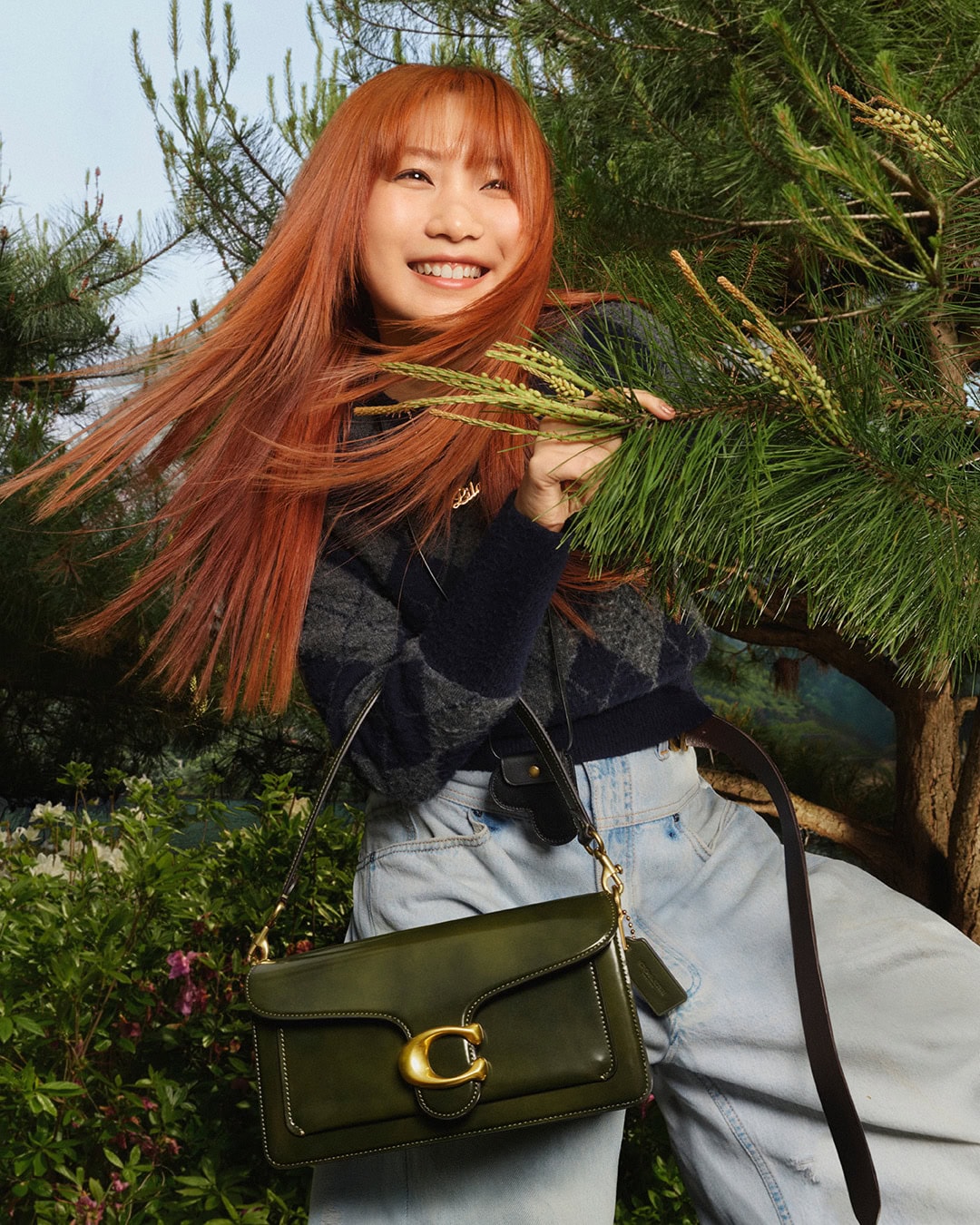 A smiling Asian woman with red hair poses outdoors with a green leather purse, wearing light-wash jeans and a patterned sweater in natural light.