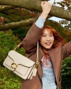 Smiling woman in a plaid blazer holding a white leather shoulder bag while gripping a tree branch outdoors