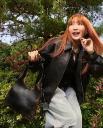 Young woman with long orange hair wearing a black leather jacket and holding a dark handbag outdoors among green foliage