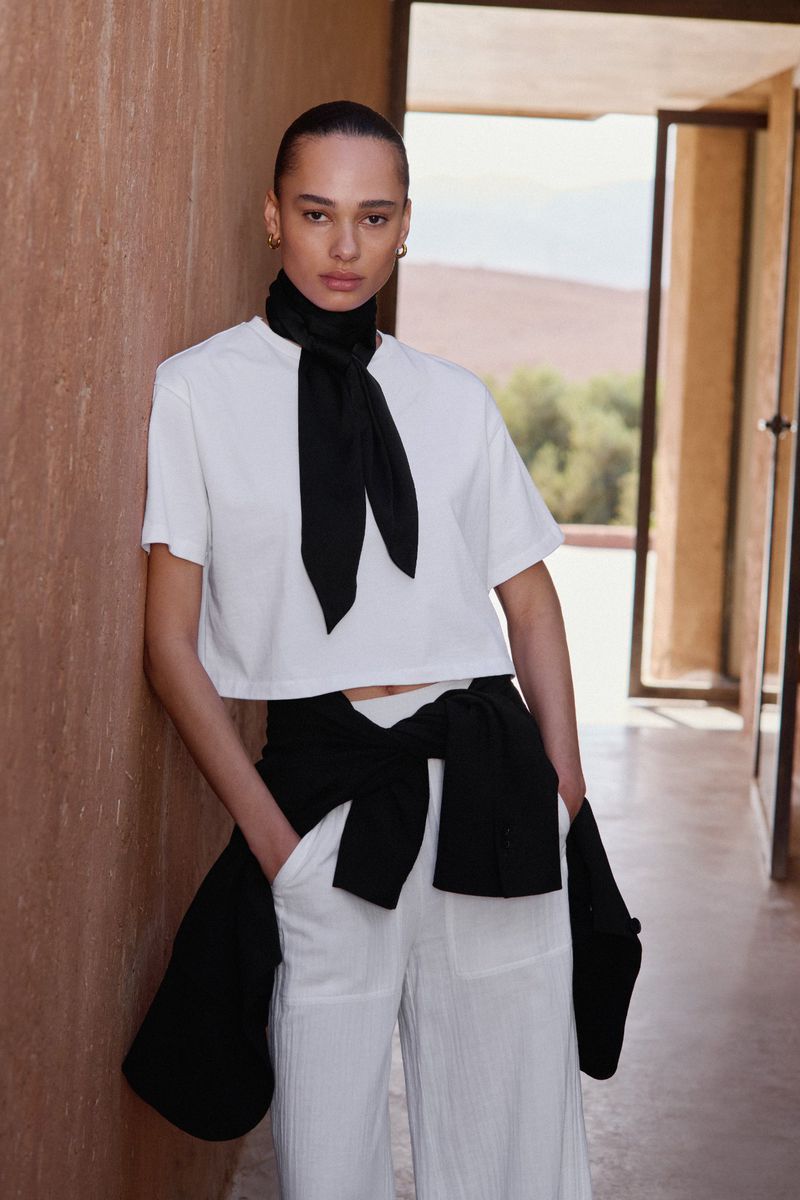 Model leaning against a textured terracotta wall wearing a white t-shirt and white trousers with a black neck scarf.