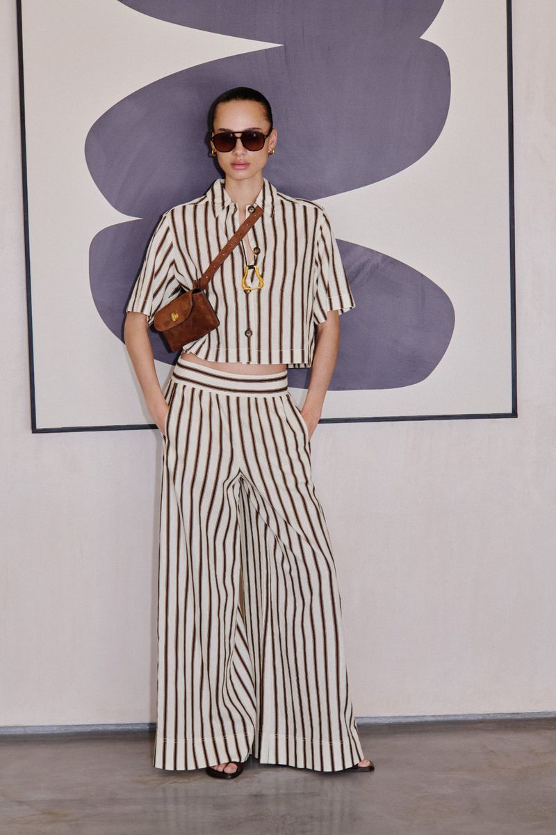 Fashionable woman in striped outfit poses against abstract art, wearing sunglasses and a crossbody bag in a studio environment.