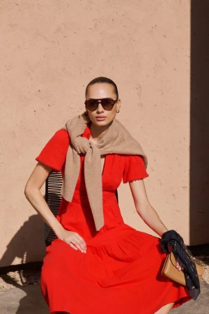 Model in red dress wearing sunglasses and beige sweater draped over shoulders against a textured wall