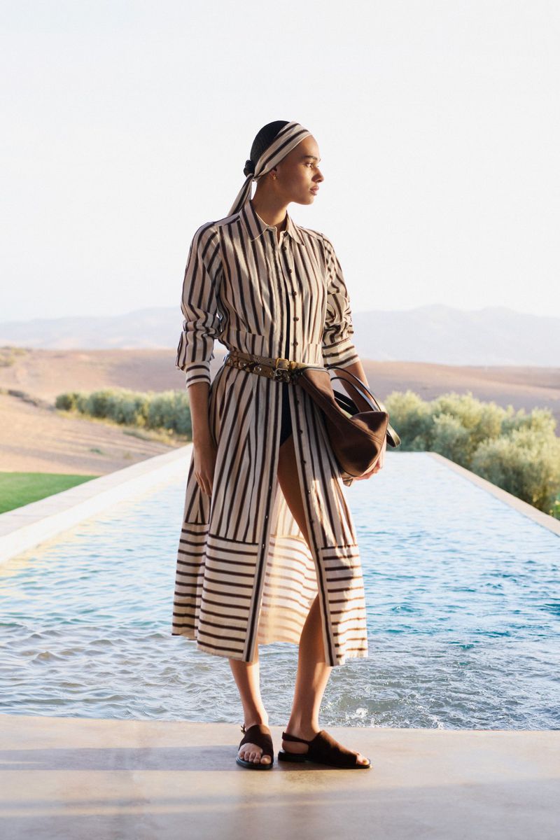 A woman wearing a long striped shirt dress and matching headscarf stands by an infinity pool with a desert landscape in the background.