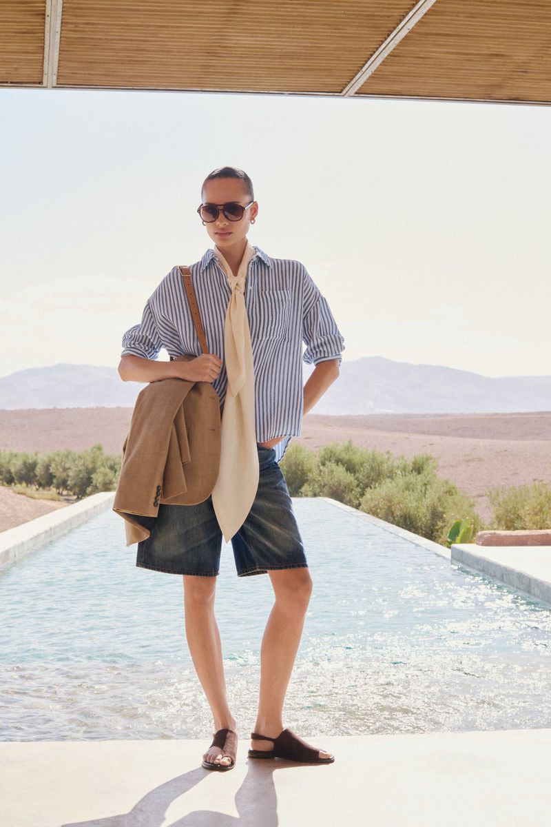 Adult female stands poolside wearing sunglasses, striped shirt, denim shorts, and brown sandals, with a jacket draped over her shoulder.