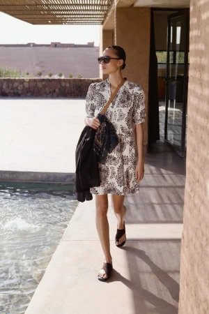 Woman in patterned dress and sandals walks by pool, carrying jacket. Sunglasses and bag. Outdoor setting with neutral tones.