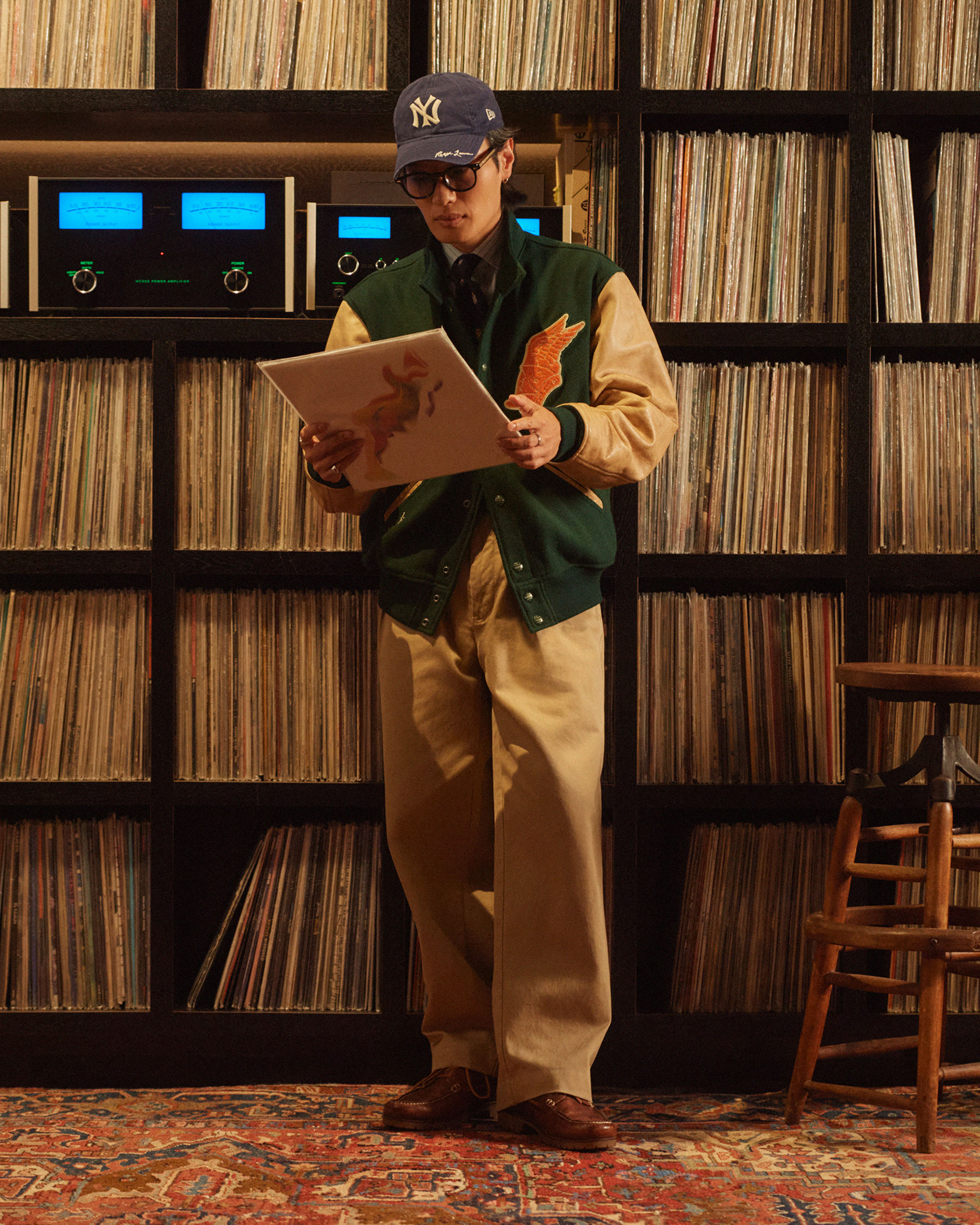 Man in baseball cap stands in a room lined with records, holding an album cover. He wears a varsity jacket and eyeglasses.