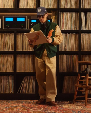 Man in baseball cap stands in a room lined with records, holding an album cover. He wears a varsity jacket and eyeglasses.