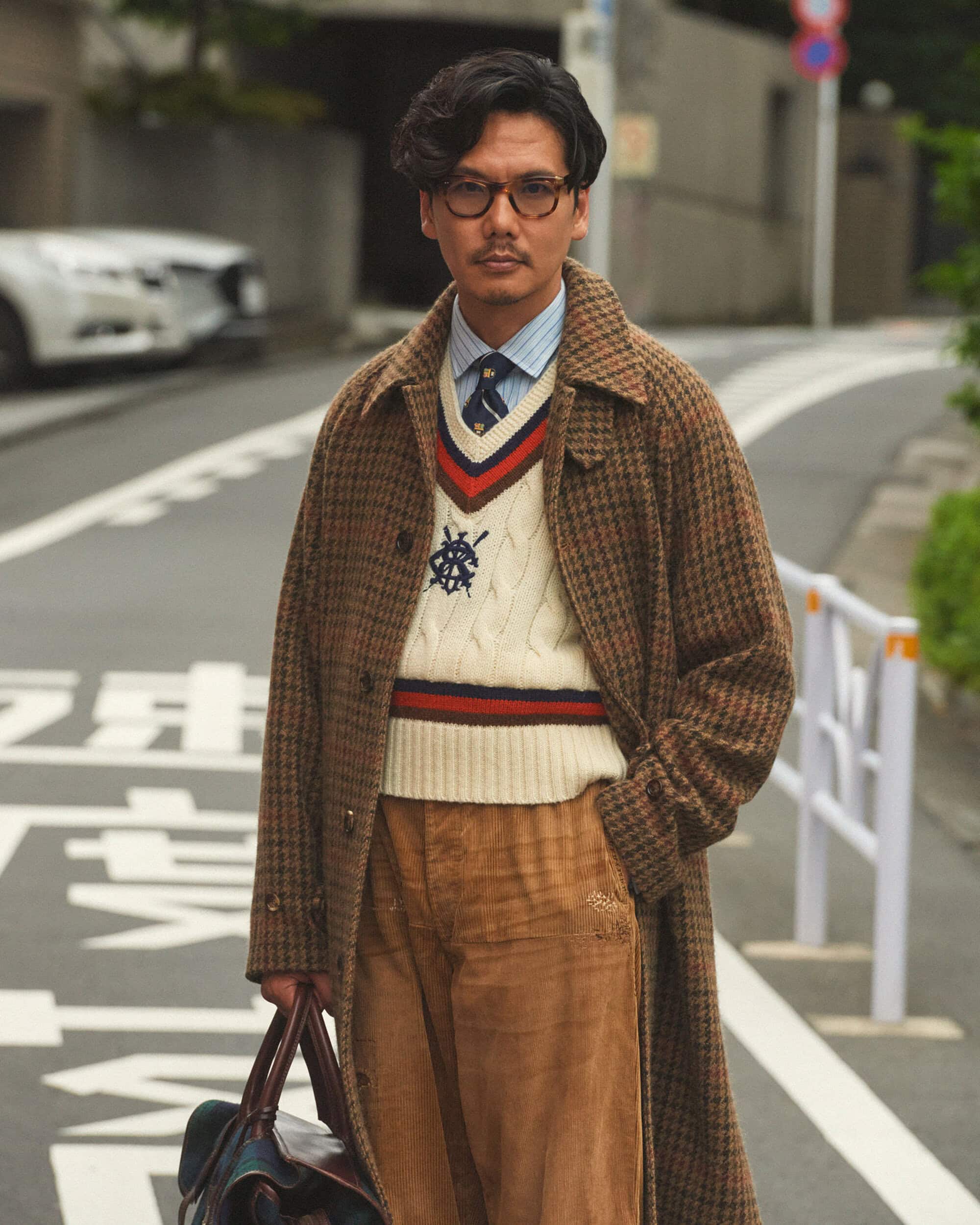 Man in a tweed coat and glasses walks on a city street carrying a bag, wearing a patterned sweater, and corduroy pants on a street.