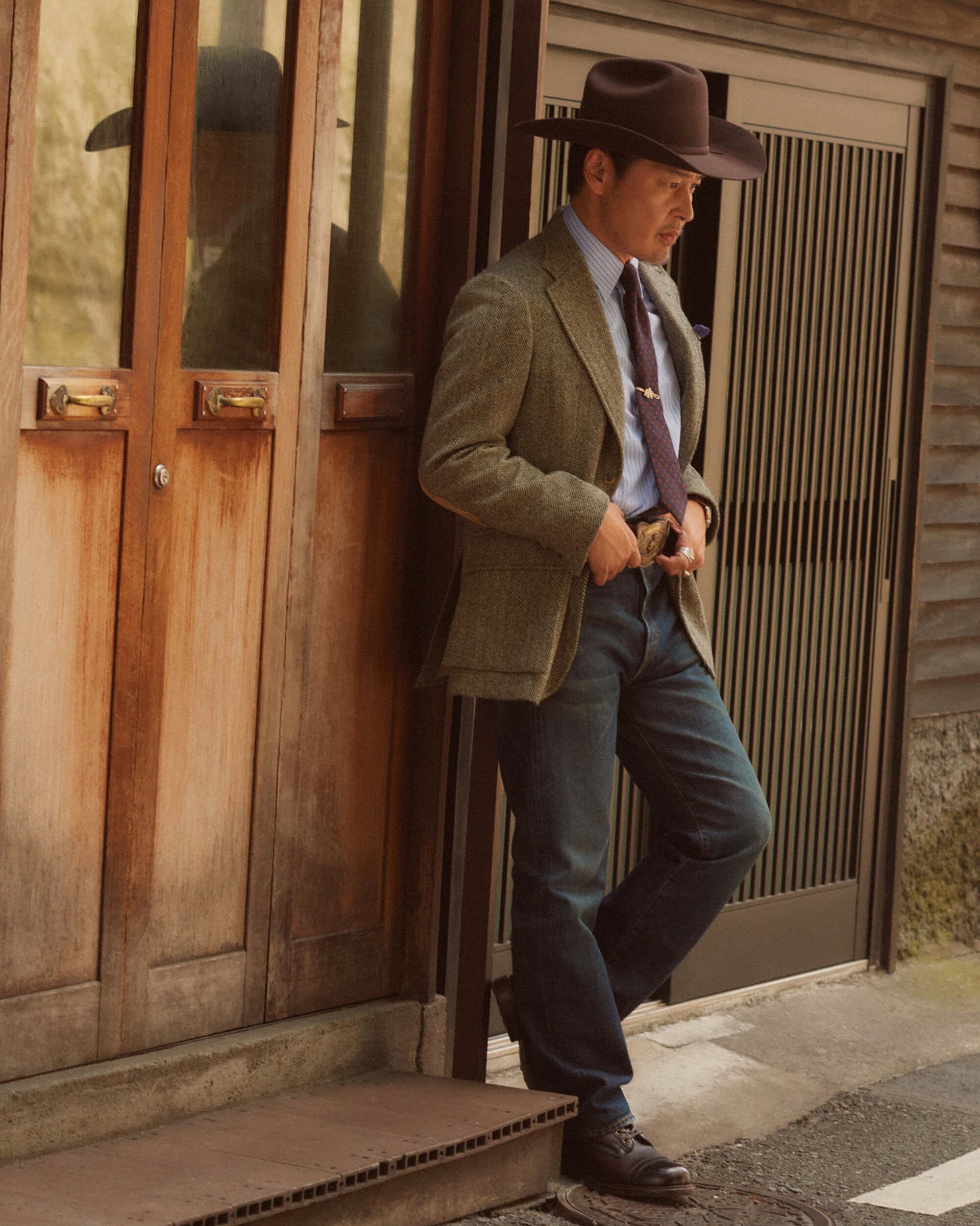 Adult man with a hat leans against a weathered wooden door outside, wearing a blazer, jeans, and boots, in an outdoor setting.