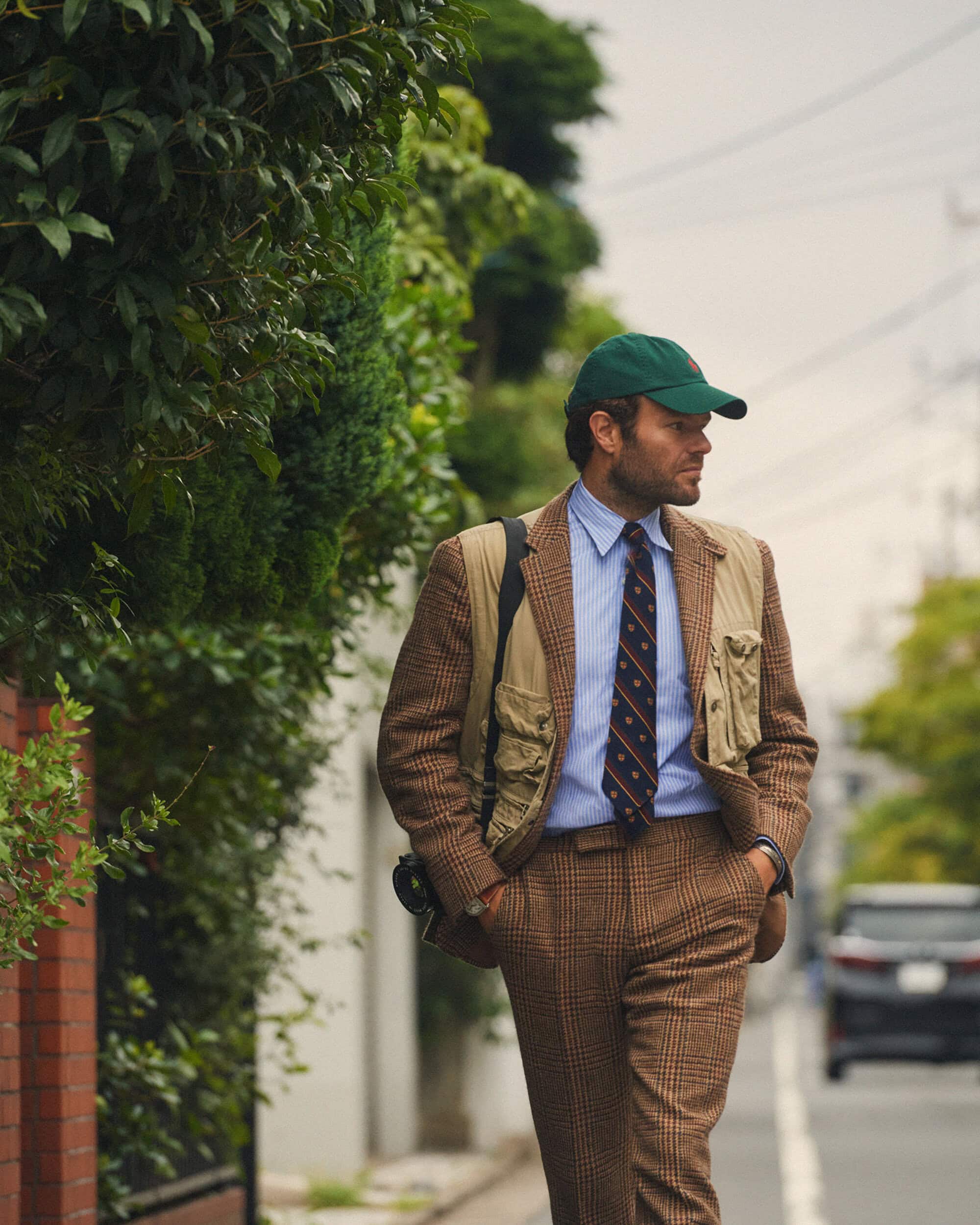 Man in brown suit with green baseball cap stands on sidewalk near a tree-lined street and a parked car under an overcast sky.