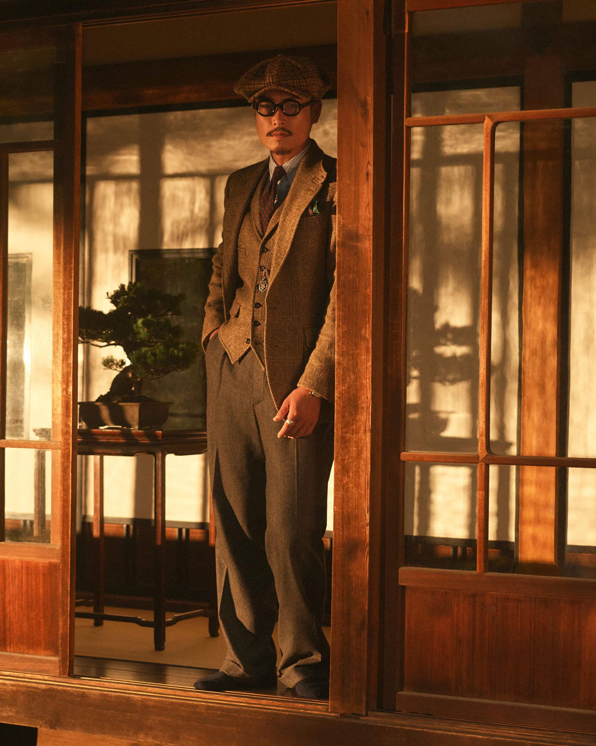 Stylish man in a tweed suit stands in a doorway with a cigarette, accented by indoor lighting and a bonsai tree.