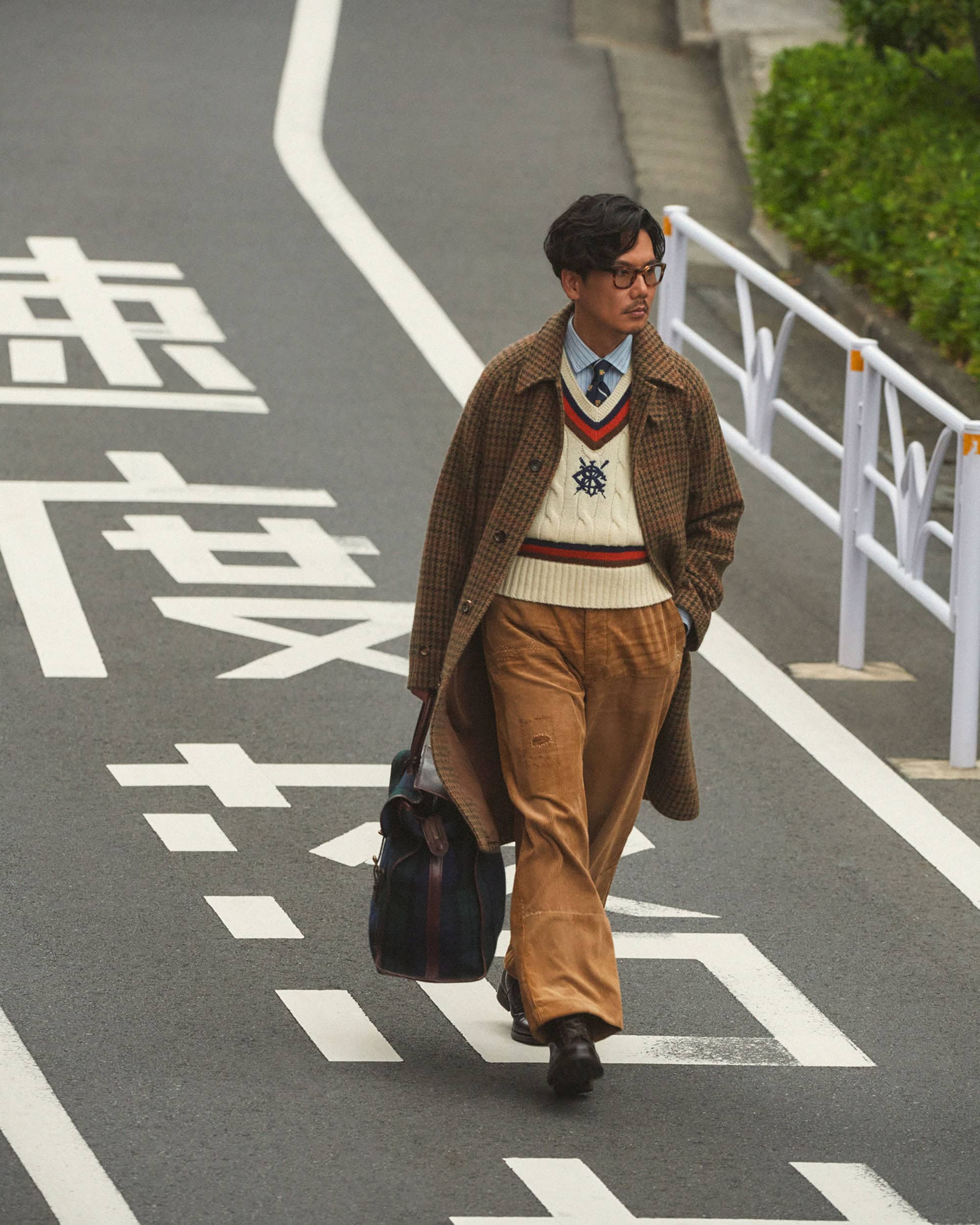 An adult man with glasses and a coat walks on a gray street with white painted text, carrying a bag and wearing brown corduroy pants.