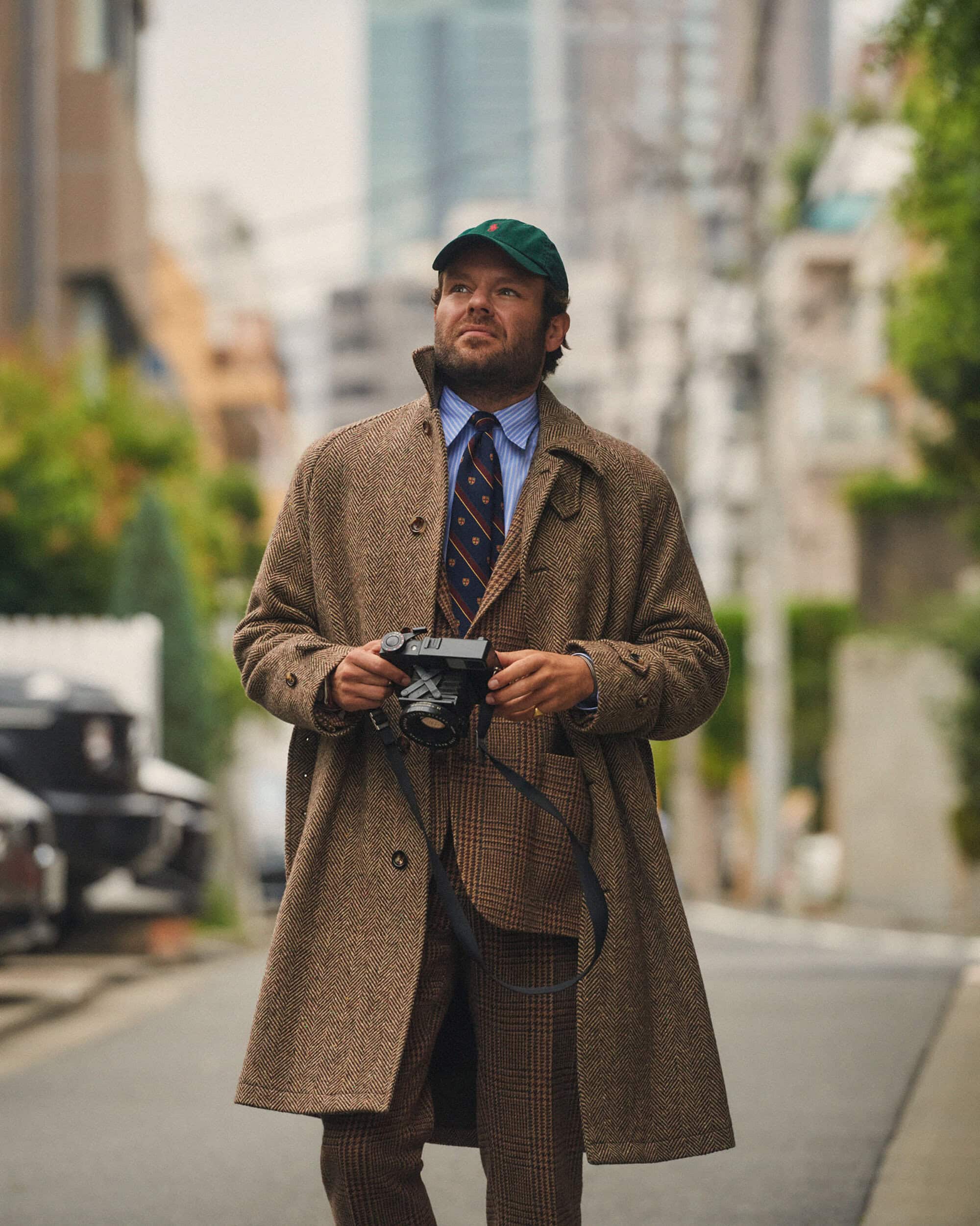 Man in tweed suit and coat stands on city street holding camera, wearing a cap and tie, with buildings and greenery in the background.