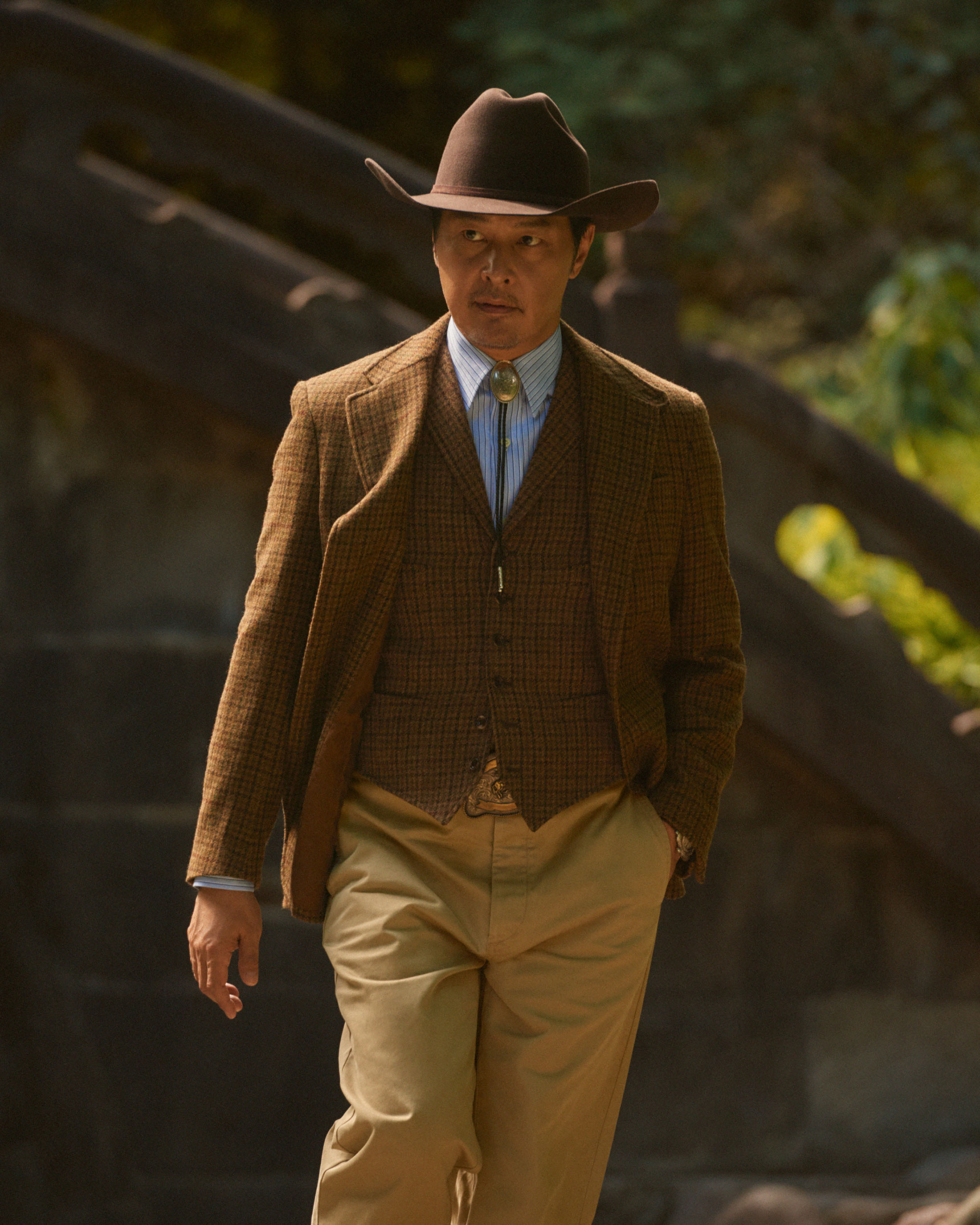 Adult man in a brown suit and cowboy hat walks in a park setting with trees and stone structures in the background, medium shot.