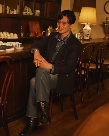Man in a blue blazer, denim shirt, and jeans sits in a warm-toned room with teacups, a samovar, and a floral lamp visible in the background.