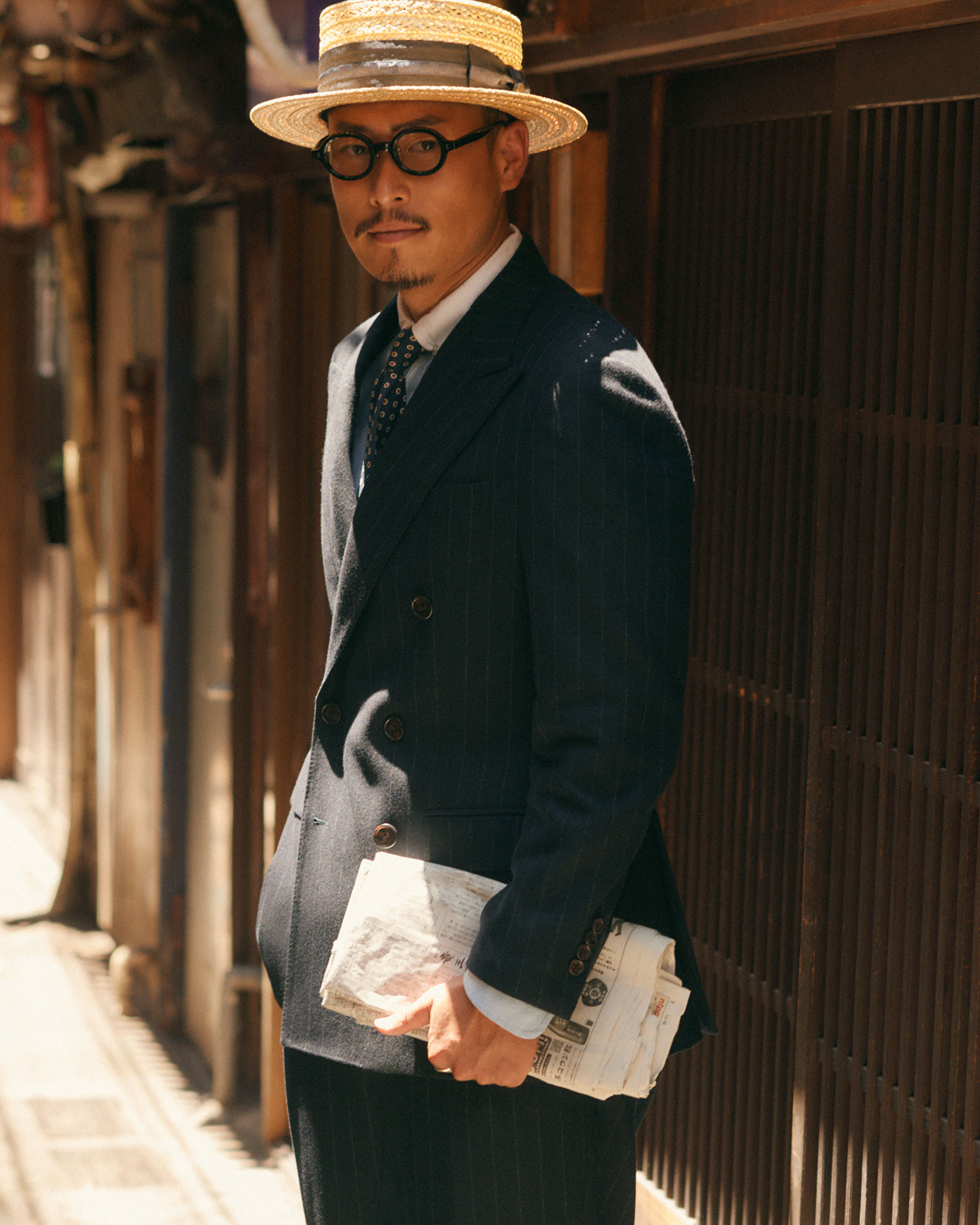 Mature adult man in a pinstripe suit and straw hat poses outdoors, holding a newspaper, in a lifestyle fashion editorial photograph.