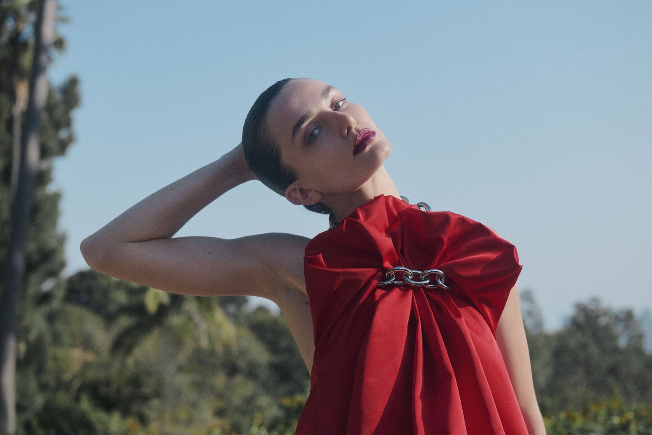 Glamorous woman in a striking red dress poses outdoors against a clear blue sky with lush green foliage in the background.