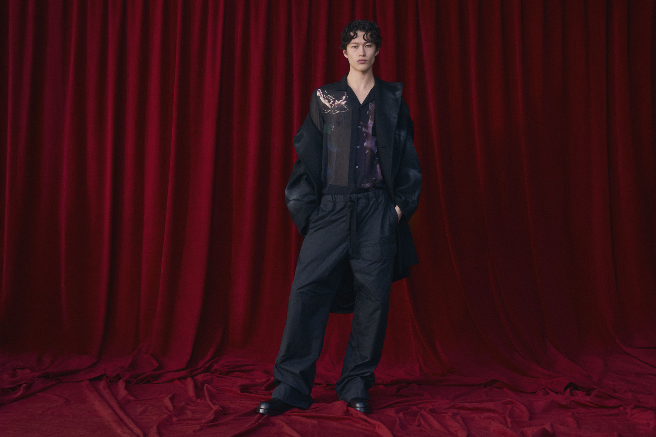 An adult fashion model stands in front of draped red curtains wearing a black coat, pants, and button-down shirt with a graphic design.