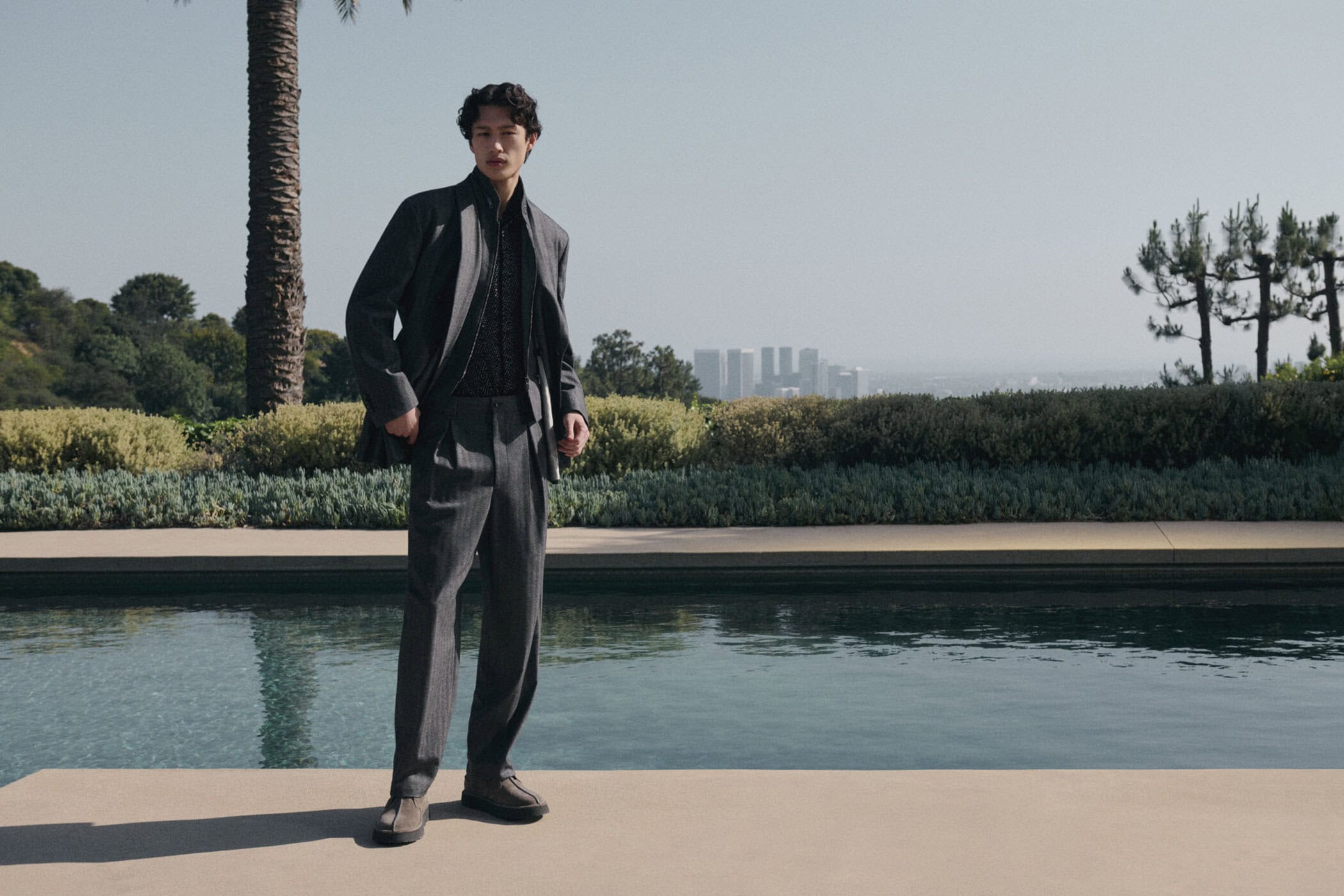 A young adult man in a grey suit poses confidently outdoors with a luxury resort pool and distant cityscape as a backdrop on a bright day.