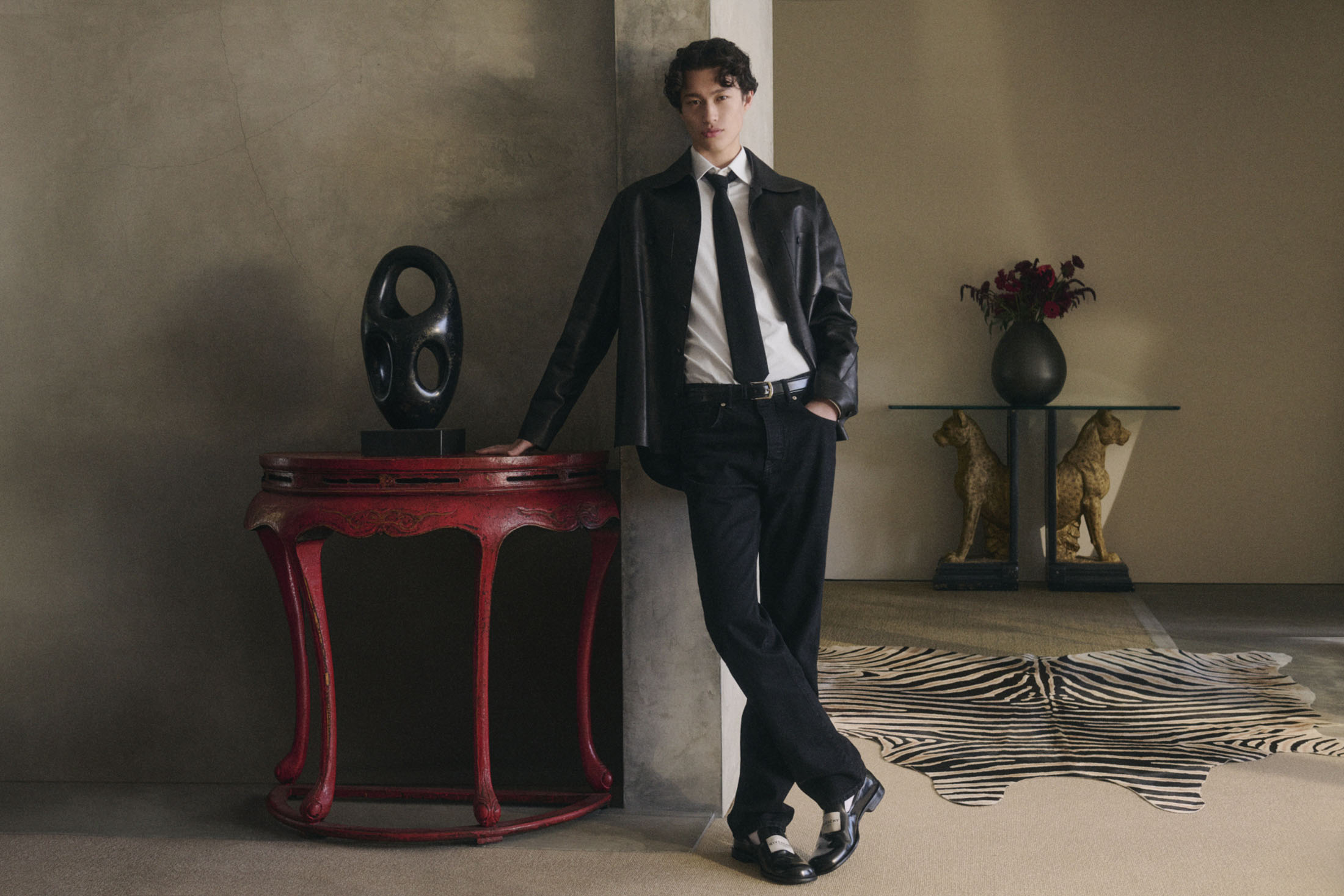 A stylish young man in a black jacket, white shirt, and black tie leans against a red table in an elegantly decorated room.