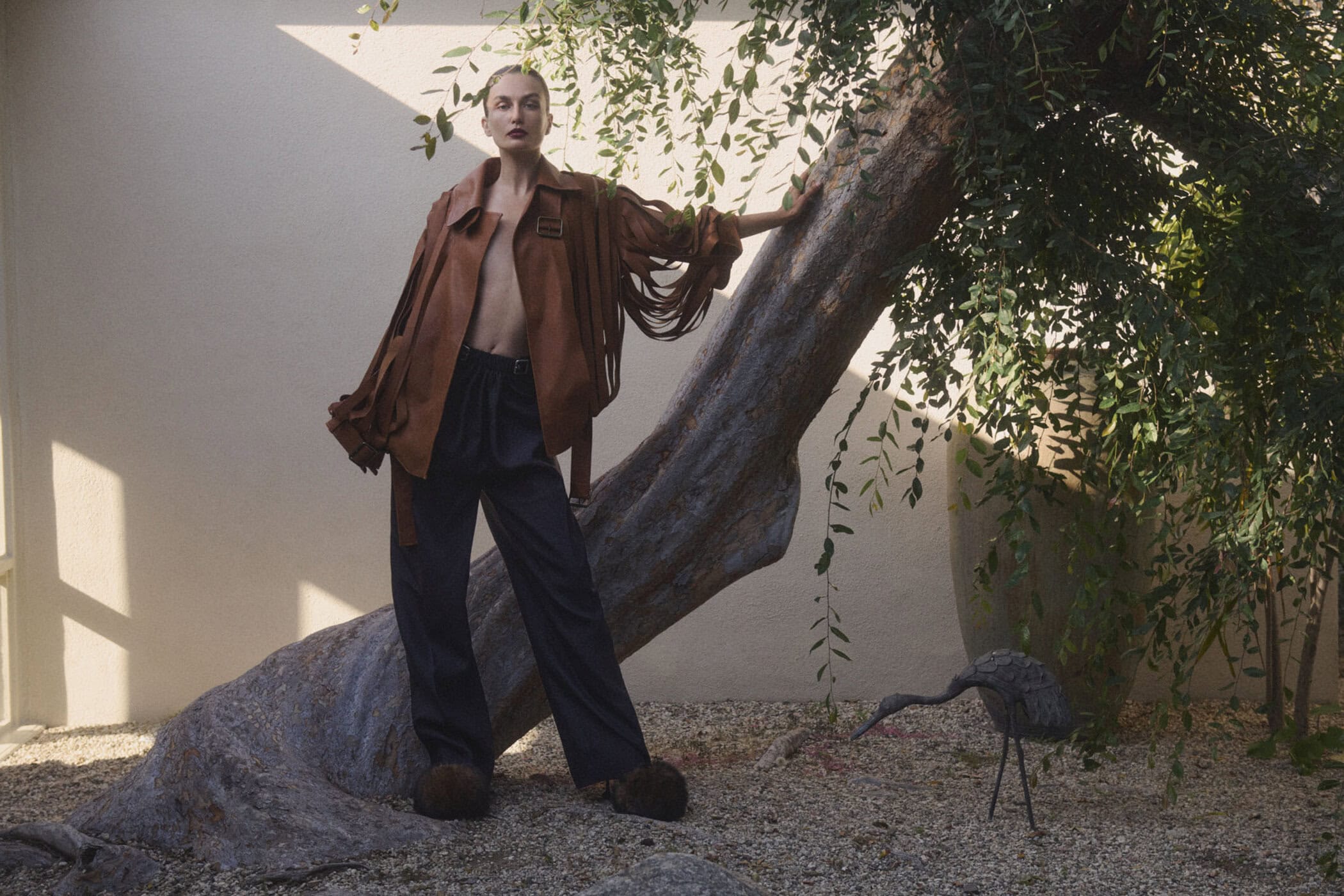 A stylish adult woman poses in a brown jacket and dark pants next to a tree outside, illuminated by soft, natural light and warm colors.