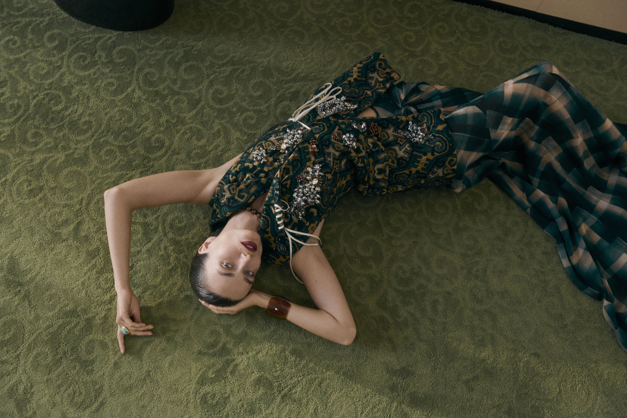 Woman in patterned dress lies on green, patterned carpet, creating a high-angle fashion portrait with soft, natural lighting and elegant pose.