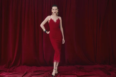 Model Andreea Diaconu in a red slip dress standing against deep red curtains for Neiman Marcus.