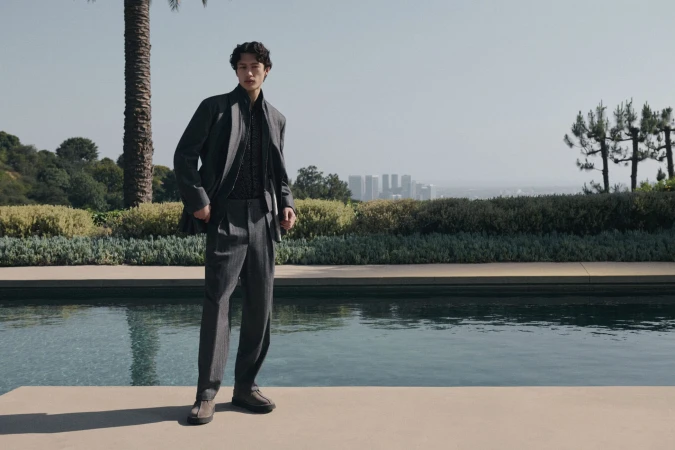 Man wearing a grey suit standing by an outdoor swimming pool with a distant city skyline in the background for Neiman Marcus