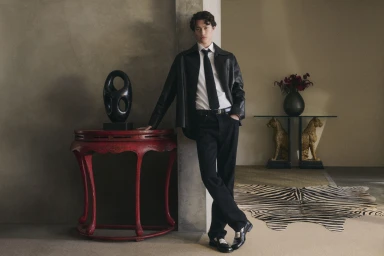 Man wearing a black leather jacket and black trousers leaning against a wall in a sophisticated interior space