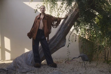 Model standing outdoors next to a leaning tree trunk wearing a brown fringe jacket and dark trousers for Neiman Marcus.