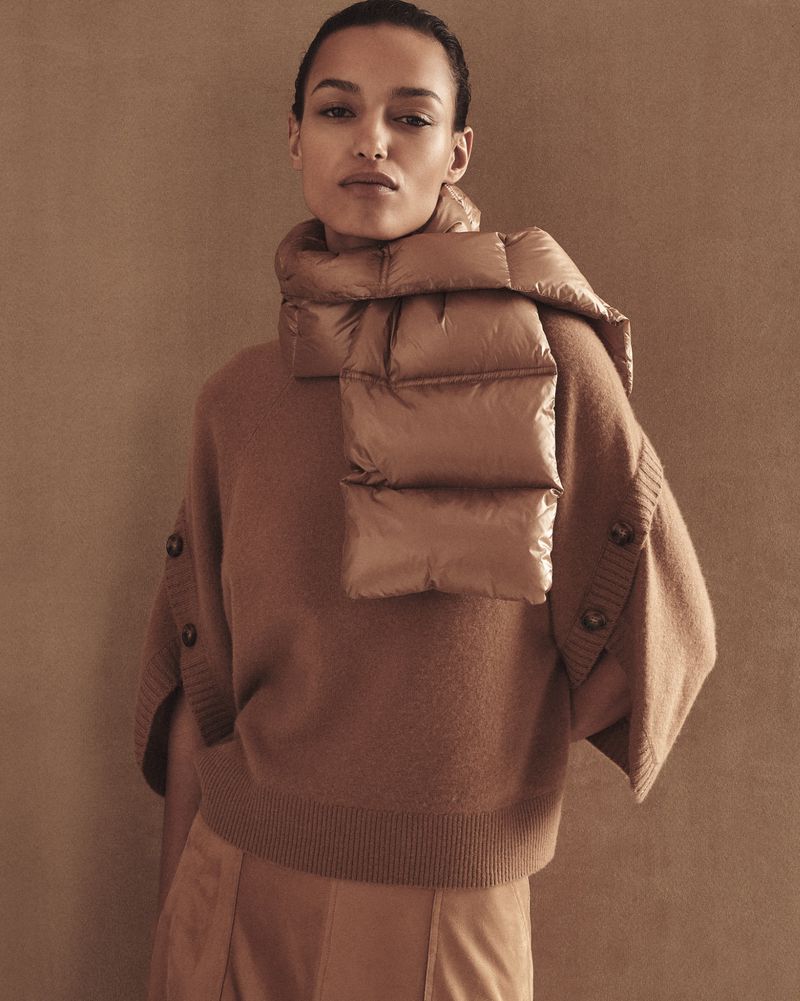 Headshot of a woman in a tan sweater and puffy scarf against a brown background in a fashion editorial style, studio lighting.