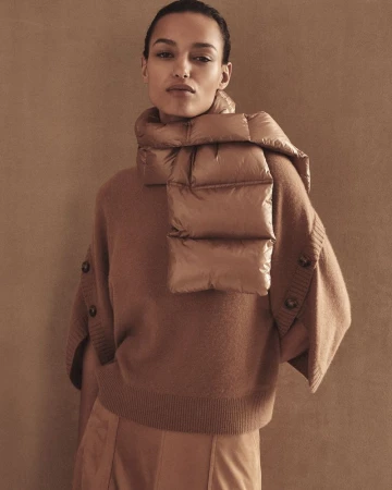 Headshot of a woman in a tan sweater and puffy scarf against a brown background in a fashion editorial style, studio lighting.