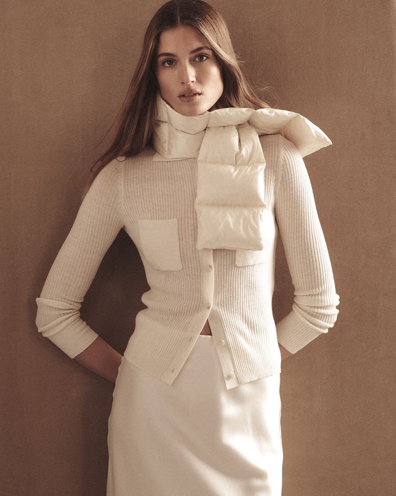 Model wearing a cream ribbed cardigan and matching puffer scarf against a warm neutral studio background.