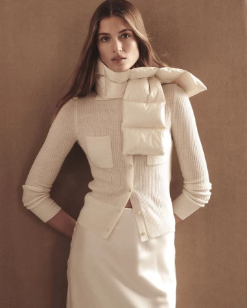 Model wearing a cream ribbed cardigan and matching puffer scarf against a warm neutral studio background.