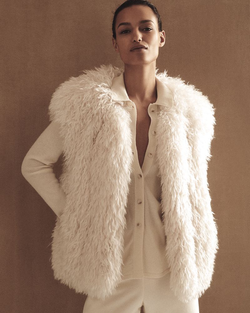 Elegant woman in white faux fur vest and matching outfit poses against a neutral backdrop in a fashion forward studio portrait.