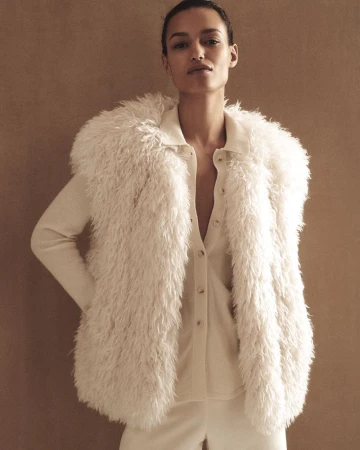 Elegant woman in white faux fur vest and matching outfit poses against a neutral backdrop in a fashion forward studio portrait.