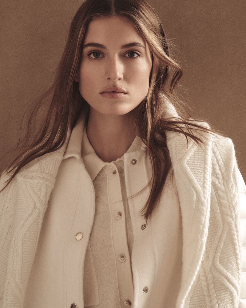 Close-up of a fair-skinned adult female model wearing a white, button-down shirt with a white cable knit sweater and coat against a tan background.