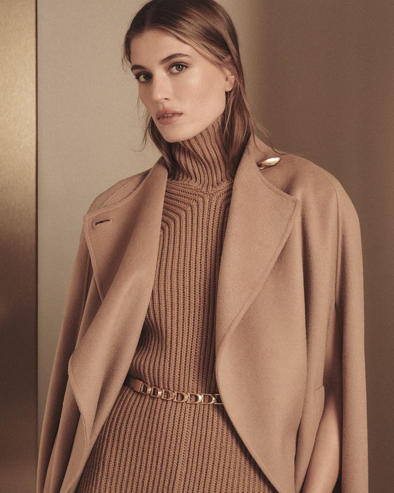 Portrait of a fair-skinned woman wearing a beige turtleneck sweater and coat, accentuated with a gold belt, in a neutral-toned indoor setting.