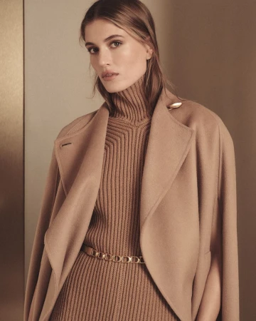 Portrait of a fair-skinned woman wearing a beige turtleneck sweater and coat, accentuated with a gold belt, in a neutral-toned indoor setting.