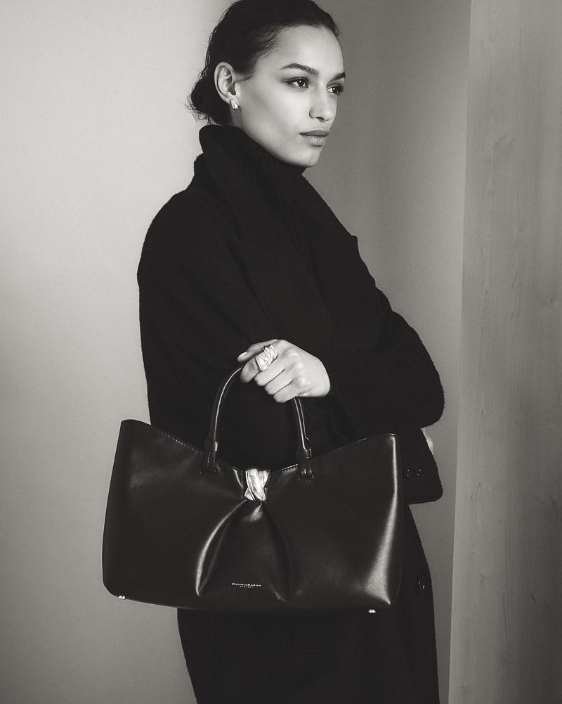 Model in black coat holding a dark leather handbag against a neutral wall for Donna Karan lookbook.