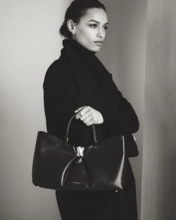 Model in black coat holding a dark leather handbag against a neutral wall for Donna Karan lookbook.