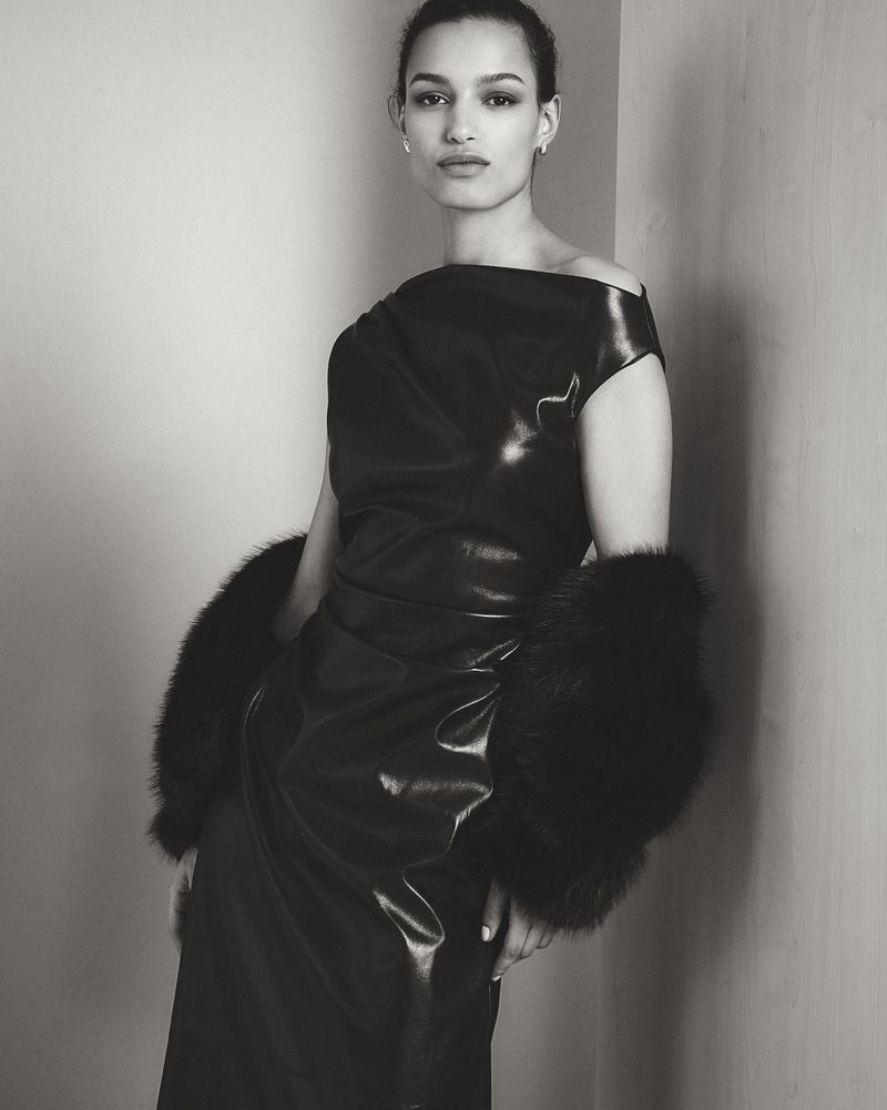 Elegant woman in a black one-shoulder dress with fur accents strikes a pose in a studio. Monochrome image captures fashion and style.