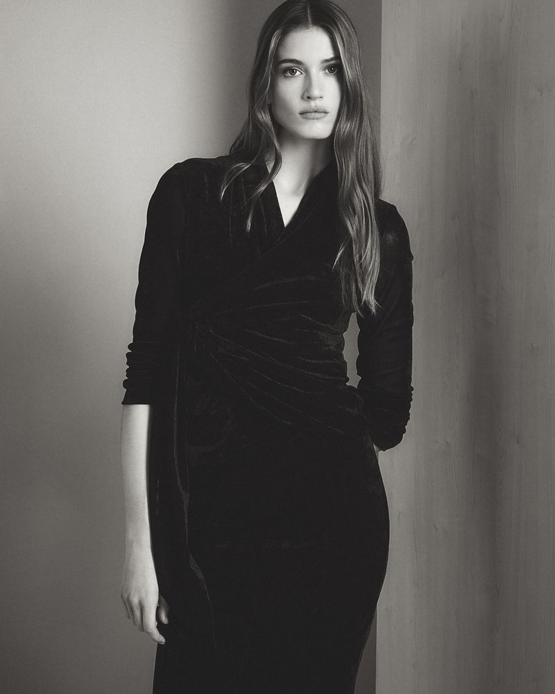 Elegant woman in a black velvet dress poses against a neutral wall. Fashion shot showcases the dress's design and fabric.