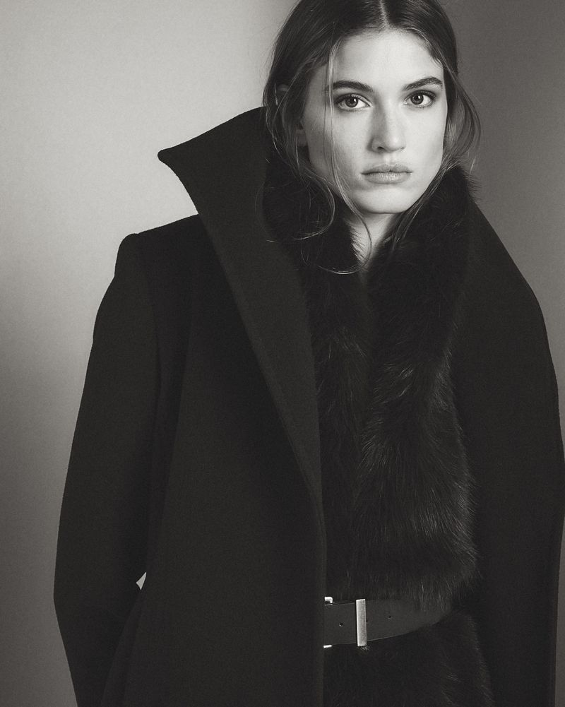 A woman wears a high-collared black coat with a faux fur trim and a black belt over a dark top, looking directly at the camera.