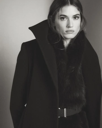 A woman wears a high-collared black coat with a faux fur trim and a black belt over a dark top, looking directly at the camera.